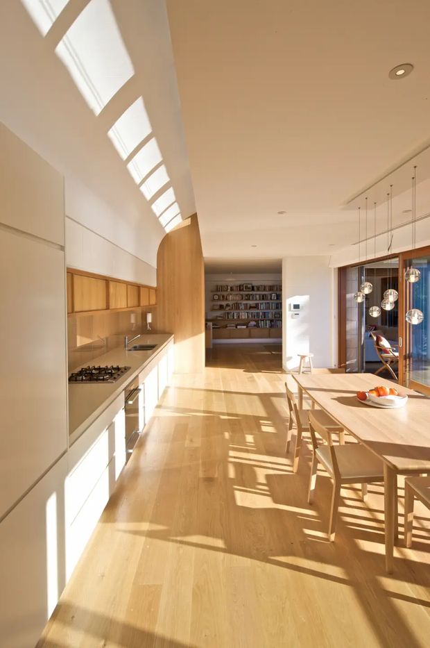 A bright, elongated open-plan kitchen and dining space with extensive pale timber cabinetry, a linear kitchen along the left wall, and a large timber dining table with matching chairs. Strong natural light enters through a series of high clerestory-style roof windows, casting dramatic geometric shadows across the timber floor. A full-height bookcase is visible at the rear, while glazed doors and spherical pendant lights define the dining area on the right.