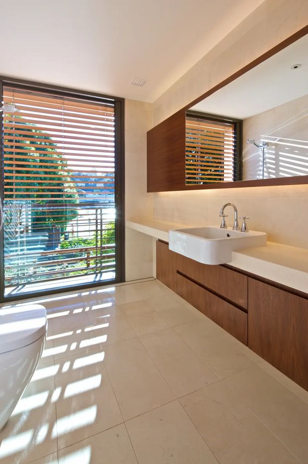 Bright contemporary bathroom with a floating timber vanity, white vessel basin, pale stone-look wall and floor tiles, and a large horizontal framed mirror. Full-height glazing with timber Venetian blinds brings in strong patterned sunlight and overlooks greenery and a balcony or exterior area.