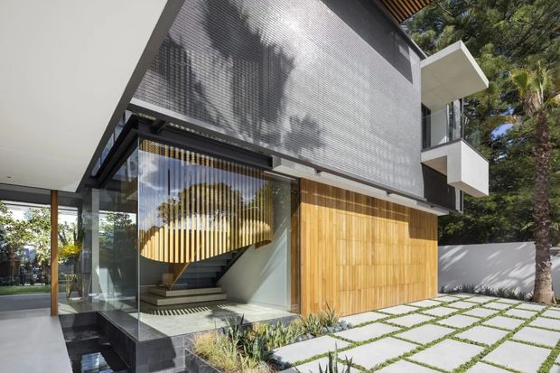Contemporary multi-level home exterior with a dark textured upper facade, extensive floor-to-ceiling glazing, warm vertical timber screening at the entry, and projecting white balconies. A sculptural curved timber feature is visible inside the glazed stair hall. The building is set within a landscaped courtyard with large pale paving slabs and planted joints.