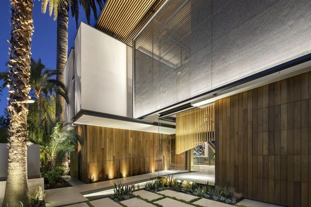 Contemporary two-storey residence viewed from the landscaped front approach, featuring warm vertical timber wall cladding, crisp white projecting volumes, extensive fine metal screening, timber slatted soffits, and integrated linear lighting. A paved path with low desert-style planting and mature palm trees leads toward the recessed entry.