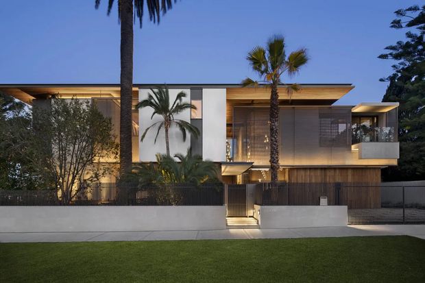 A contemporary two-storey residential facade photographed at dusk, with broad horizontal roof overhangs, warm timber soffits and cladding, large glazed openings, screened upper-level spaces, and a restrained white and grey street-facing boundary wall. Palm trees and a manicured lawn frame the house, with warm interior and entry lighting creating a refined evening presentation.