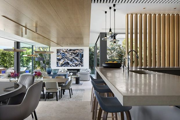 Open-plan contemporary kitchen, dining, and living area with a large pale stone island and integrated seating, dark blue bar stools, pendant lights, timber cabinetry, and a vertical timber-screen feature. Floor-to-ceiling glazing brings in abundant natural light and overlooks greenery. A dining table and lounge seating are visible beyond the kitchen.