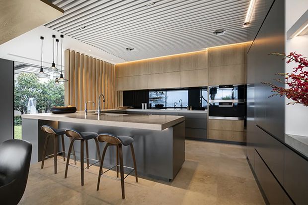 A sophisticated contemporary kitchen with a large central island and four timber-and-leather bar stools. The space features warm stone-look flooring, dark built-in cabinetry, pale timber cabinetry and wall panelling, a black splashback, integrated appliances, pendant lighting, and a striking linear slatted ceiling with concealed illumination. Full-height glazing connects the kitchen to a leafy outdoor area.