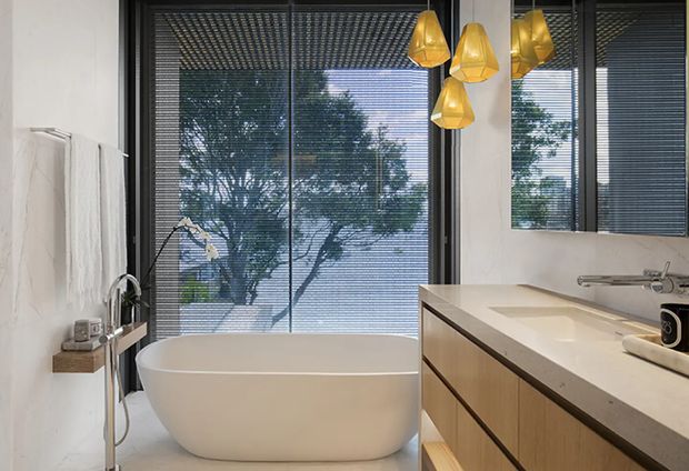 Contemporary bathroom featuring a freestanding white bathtub positioned in front of large dark-framed windows with horizontal blinds. A floating timber vanity with a pale stone benchtop and integrated basin occupies the right side, while a wall-mounted shower fixture and small ledge appear on the left. Warm sculptural pendant lights hang above the vanity, and trees are visible outside through the glazing.
