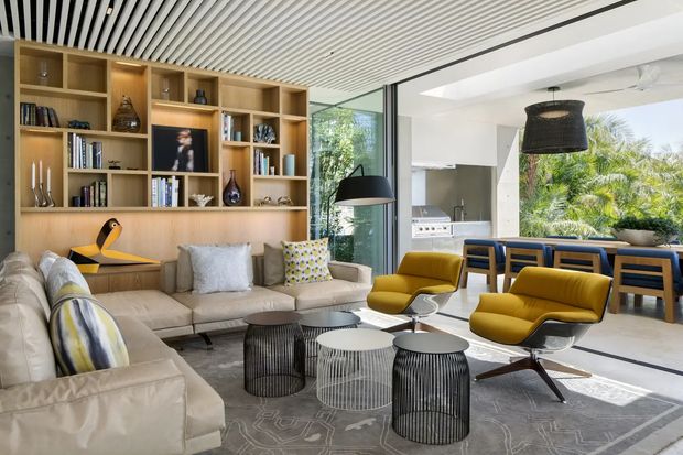 Open-plan contemporary living and dining area with a large built-in timber shelving wall, sectional sofa, sculptural lounge chairs, nesting coffee tables, and a dining table beside the kitchen. Full-height glazing connects the interior to lush greenery outside. A linear slatted ceiling and restrained neutral palette are balanced by mustard-yellow seating and patterned cushions.