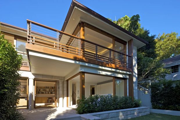 Exterior view of a contemporary two-storey residence with a prominent upper-level balcony, slim metal balustrades, warm timber accents, extensive glazing, and a low-pitched roof. The ground level includes a sheltered open area and visible built-in shelving, while landscaped planting and a stone-edged garden frame the facade.