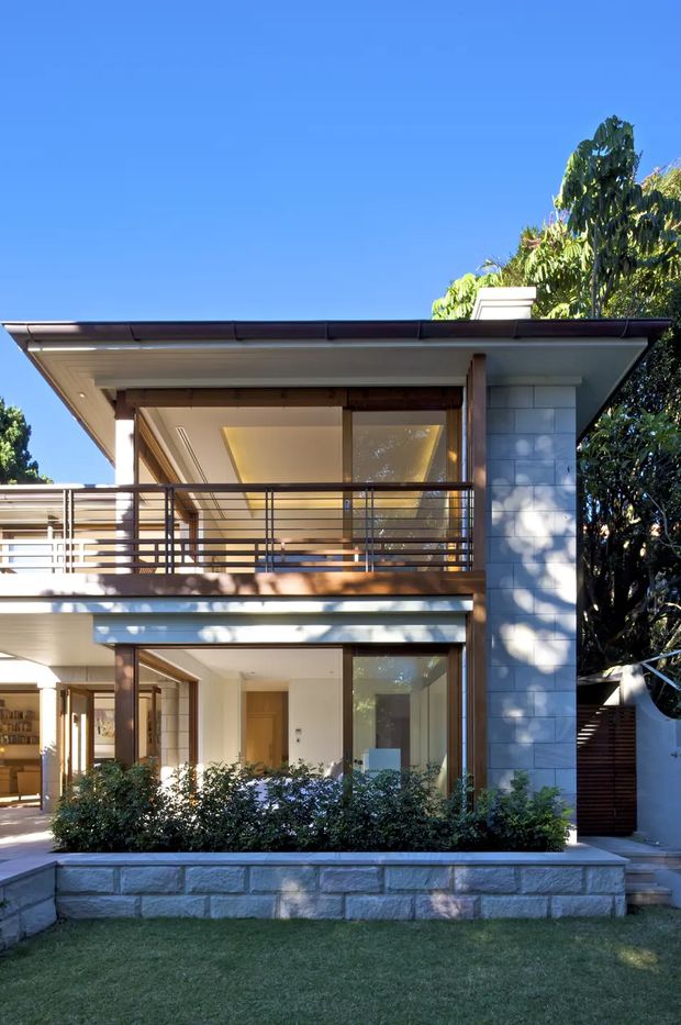 Front exterior view of a contemporary two-storey residence with a broad low-pitched roof, open upper balcony, horizontal metal balustrade, extensive glazing, timber structural accents, and pale rectangular masonry cladding. A landscaped garden bed and low stone retaining wall sit in front of the house.