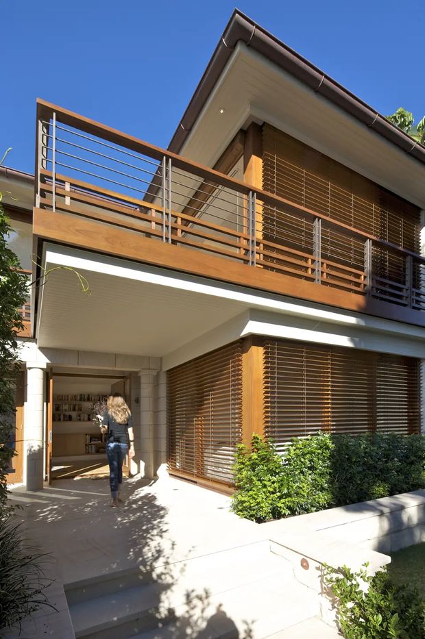 Street-level view of a two-storey contemporary residence with a projecting upper balcony, warm timber privacy louvres and balcony screening, white rendered walls and soffits, and slim metal balustrades. The entry is recessed beneath the balcony, with paved steps, low landscaping, and strong sunlight creating a bright suburban facade.