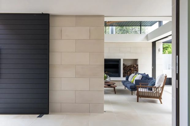 A bright contemporary living room viewed through a wide opening, with pale large-format stone or porcelain wall and floor finishes, a dark horizontal-panel feature wall, built-in fireplace and wood storage, blue sectional seating, and a timber lounge chair. High-level glazing and clerestory windows bring in abundant natural light.