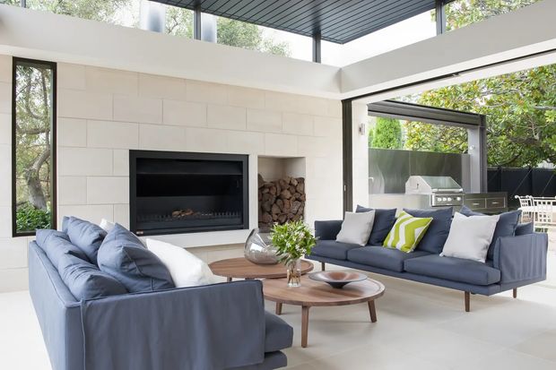 Bright, open-plan living room with two blue-grey sofas arranged around layered round timber coffee tables. A large black built-in fireplace is set into a pale tiled feature wall with a recessed firewood niche. Floor-to-ceiling glazing and high clerestory windows bring in abundant natural light, while a covered outdoor kitchen is visible beyond the open doors. The space has a clean contemporary feel with pale surfaces, dark metal framing, and bold green-and-white cushions.