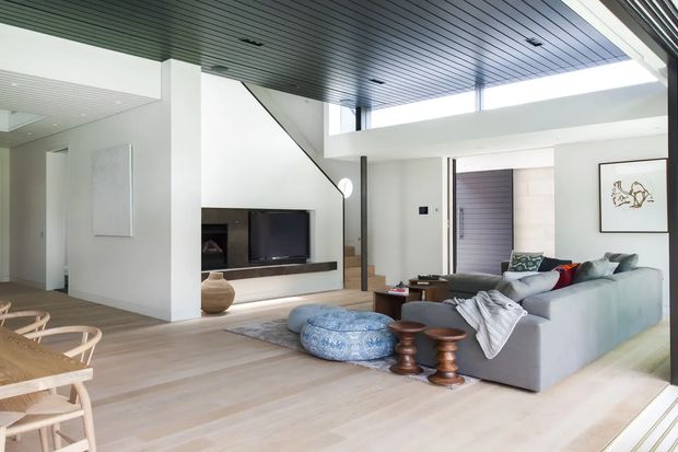 Bright, open-plan contemporary living space with a high ceiling and mezzanine-like upper level. The room features pale timber flooring, white walls, a dark slatted ceiling, a wall-mounted television with low dark joinery, and a large grey sectional sofa. A dining area is visible in the foreground, with a staircase and entry zone in the background. Large glazing brings in abundant natural light.