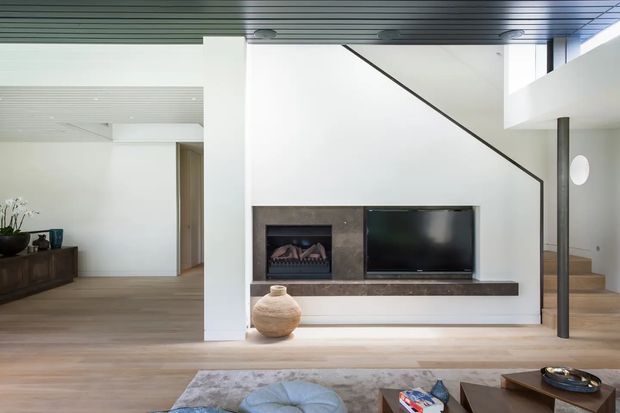 Contemporary open-plan living area with a minimalist built-in media wall and fireplace, pale timber flooring, white walls, and a stairway rising behind a slim dark structural post. A partially visible dining or entry area extends to the left, with a low timber console and sparse decor. The space has clean linear detailing, a partially double-height feel, and restrained neutral styling.