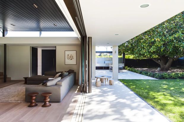 A contemporary indoor-outdoor living area with a large sectional sofa, low coffee table and sculptural stools opening onto a broad covered terrace. Full-height sliding glass doors are retracted into a dark timber-lined wall, creating a seamless connection to the manicured lawn and mature garden beyond. The interior has pale timber flooring, neutral walls, dark linear ceiling panelling and abundant natural light.