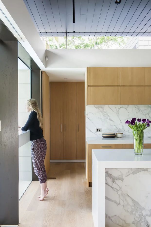 Bright contemporary kitchen with warm timber cabinetry, a white marble-look splashback and island, pale timber flooring, and extensive glazing. A high slatted ceiling incorporates a long skylight or clerestory opening, bringing in abundant natural light. The island features a waterfall-style stone end panel, while a full-height timber storage wall is visible beyond.