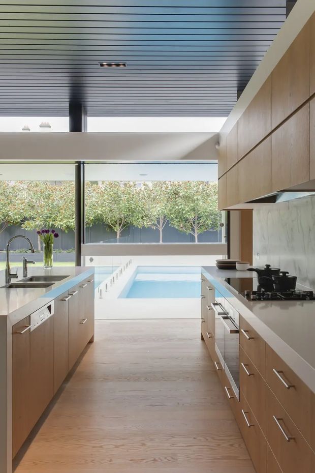 A bright contemporary kitchen with a long galley-style layout, light timber cabinetry, pale stone benchtops, and a marble-look splashback. Stainless steel appliances and a deep sink line the counters. Full-height glazing opens the kitchen toward a courtyard with a rectangular swimming pool, while a slatted ceiling and clerestory-like opening bring in abundant daylight.