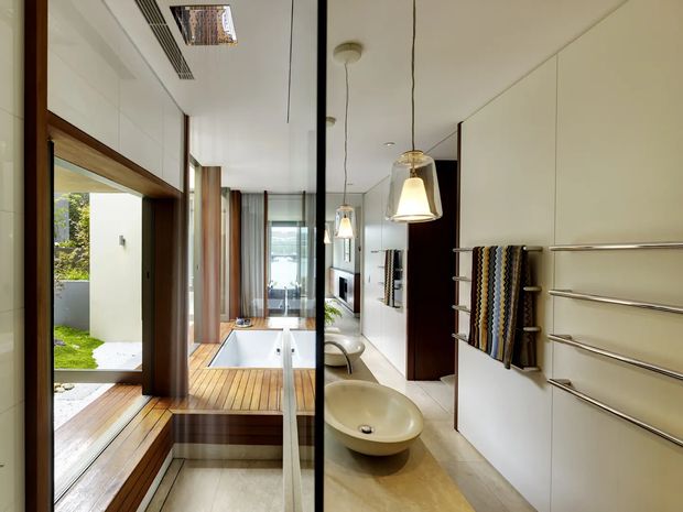 Contemporary bathroom with a long, narrow layout, featuring a freestanding oval basin on a light stone or timber-toned surface, a walk-in shower or wet-room area, extensive mirrored cabinetry, timber-framed glazing, and warm timber decking beside an outdoor courtyard. Pendant lights and horizontal heated towel rails add refined functional detailing.