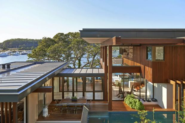 Exterior architectural view of a multi-level contemporary residence overlooking a waterfront inlet. The home features broad floor-to-ceiling glazing, warm vertical timber cladding, exposed timber posts and beams, layered flat and low-pitched roof forms, and a compact swimming pool adjoining a timber deck and courtyard-like outdoor space. Boats and a tree-covered shoreline are visible beyond the property.