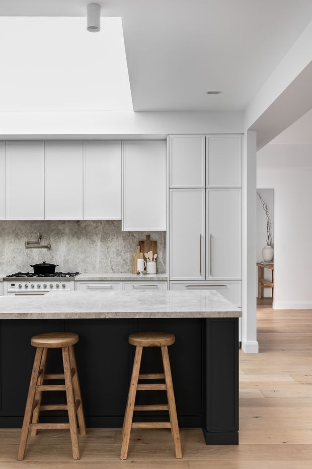 Bright, refined kitchen featuring white Shaker-style cabinetry, a large pale stone island with a black paneled base, two timber stools, a textured stone backsplash, and integrated tall storage. Light timber flooring continues into the adjoining space, while recessed and cylindrical ceiling lighting create a clean, contemporary look.