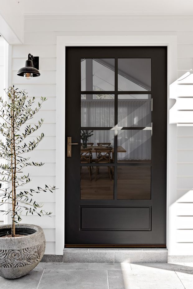 A close-up exterior view of a black, multi-pane glazed entry door set within a white weatherboard facade. The door has brass-toned hardware and a lower recessed panel, with a textured stone-paved threshold and a large decorative planter containing a slender olive tree. Reflections through the glazing reveal a dining table and curtains inside.