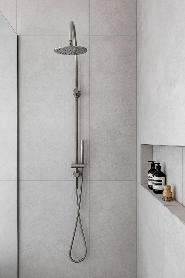 Minimalist walk-in shower featuring large-format light grey tiles, a brushed metal rainfall shower and handheld rail set, frameless glass screen, and a recessed tiled niche with dark toiletry bottles and a wooden brush.