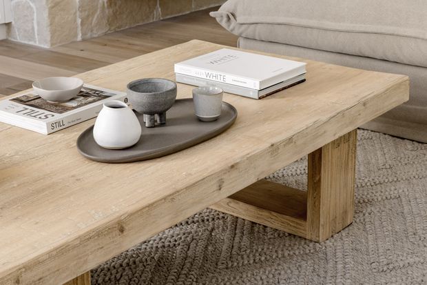Close-up interior scene featuring a large rustic light timber coffee table with chunky legs and visible natural grain. A dark ceramic tray holds a white vase, grey bowl, and cup, while books and a neutral upholstered sofa sit behind it. The room includes a textured stone wall, warm timber flooring, and a chunky woven grey rug, creating a calm, warm, natural aesthetic.