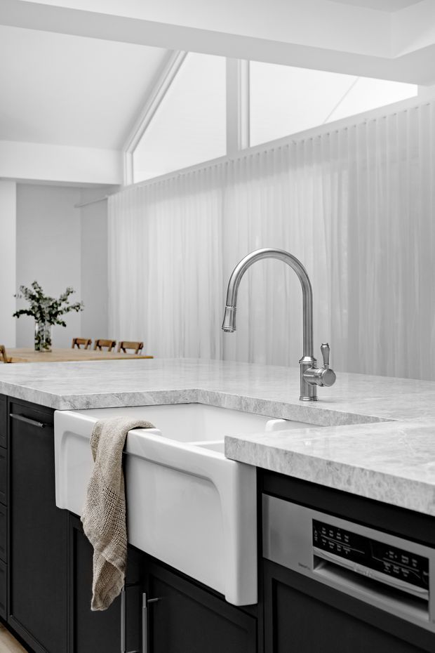 Bright, refined kitchen interior featuring a large white farmhouse apron-front sink, tall polished metal gooseneck faucet, pale veined stone benchtops, and dark cabinetry. A vertically ribbed translucent partition and high clerestory glazing bring diffused natural light into the space, with a dining area visible beyond.