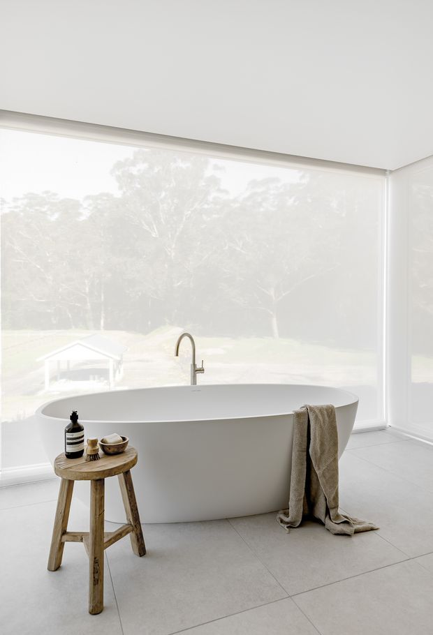 Bright, minimalist bathroom featuring a freestanding white oval bathtub positioned in front of expansive corner glazing with translucent roller blinds and views of trees outside. A slim brushed-metal tap rises from behind the tub. Large light-grey floor tiles, white walls and ceiling, and a rustic wooden stool with bath accessories and a draped neutral towel complete the calm, refined space.