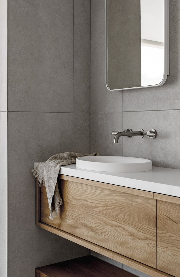 Close-up view of a contemporary bathroom vanity. Large grey concrete-look wall tiles frame a wall-mounted oak vanity with integrated drawers, topped by a white countertop and round white vessel basin. A brushed metal wall-mounted tap and rounded-rectangle mirror complete the restrained, minimalist composition.