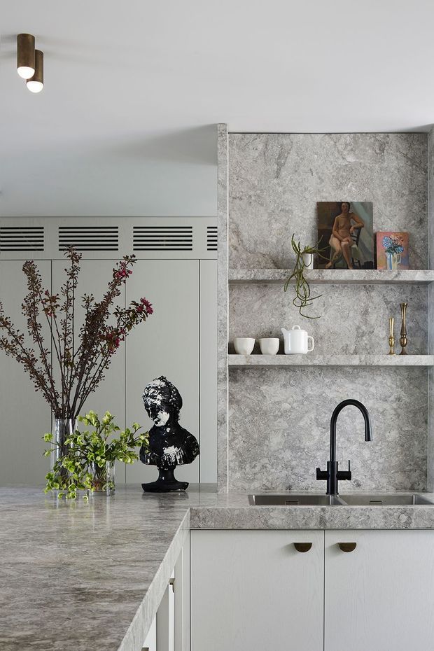 A refined contemporary kitchen with a large pale grey stone island and matching stone splashback. White flat-panel cabinetry has small dark curved pulls, while open stone shelves display ceramics, artwork, brass candlesticks and a trailing plant. A black gooseneck mixer tap, integrated sink, sculptural black bust and tall floral arrangement add contrast. The room has a light neutral palette and understated decorative styling.