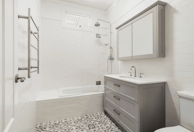 Bright, compact bathroom with white horizontal wall tiles, a bathtub and glass shower screen, chrome fixtures, heated towel rail, grey vanity cabinetry with a white benchtop, mirrored wall cabinet, and patterned encaustic-style floor tiles. The space has a clean, refined traditional-contemporary appearance.