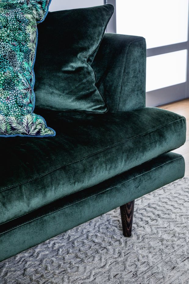Close-up interior detail of a deep emerald-green velvet upholstered armchair or sofa with a patterned botanical cushion, tapered dark timber leg, pale textured rug, timber floor, and a large dark-framed window in the background.