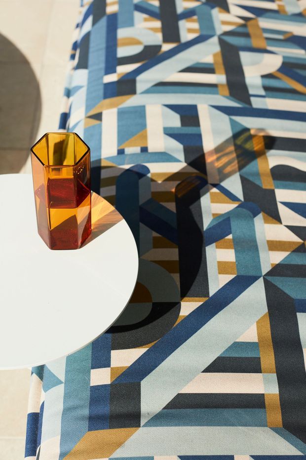 Close-up interior styling vignette featuring a bold geometric upholstered or textile surface in blue, teal, cream, black, and ochre. A small faceted amber glass tumbler sits on a round white table, with strong sunlight creating pronounced shadows across the patterned fabric. The image emphasises colour, pattern, and decorative furnishing rather than a complete room.