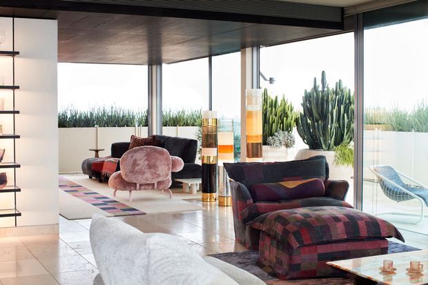 A bright, stylish mid-century-inspired living room with floor-to-ceiling glazing, a dark wood-toned ceiling, polished pale tiled flooring, and expansive views to a landscaped terrace. The room contains sculptural lounge chairs, a large patterned sectional with ottoman, a white curved sofa, open shelving, and colourful cylindrical decorative screens or shelving elements. Tall cacti and grasses are visible outside.
