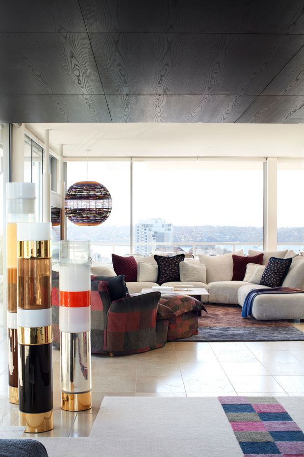 Bright, high-rise living room with floor-to-ceiling glazing, a large cream sectional sofa, patterned lounge chairs and ottomans, and colorful sculptural pendant and floor lighting. The room has a dark timber-lined ceiling, pale tiled flooring, and a contemporary, artful furnishings palette with views over a city landscape.