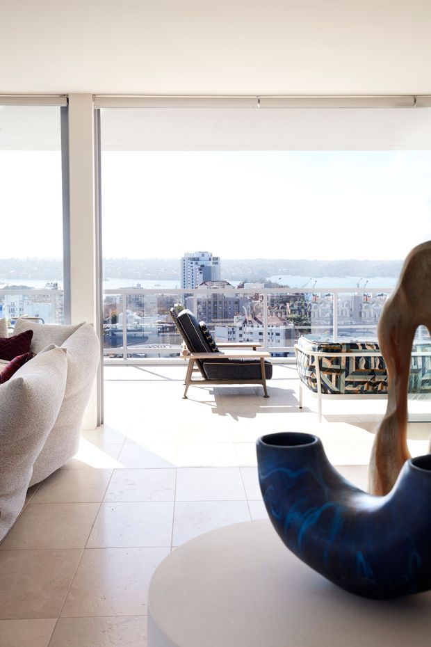 Bright contemporary living room opening onto a balcony or terrace, with expansive floor-to-ceiling glazing and panoramic views over a dense coastal city and water beyond. The room features pale tiled flooring, a light upholstered sofa, a lounge chair, patterned outdoor-style seating, roller blinds, and a sculptural dark blue ceramic vase on a round table. The overall atmosphere is airy, refined, and sunlit.