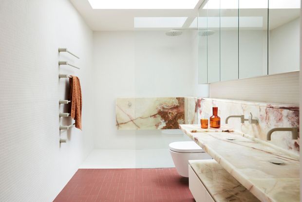 Bright contemporary bathroom with a long double vanity clad in veined natural stone, wall-mounted mirrored cabinetry, a white toilet, glass shower enclosure, skylights, and a red-toned tiled floor. A heated towel rail with a rust-coloured towel is mounted on the textured white wall.