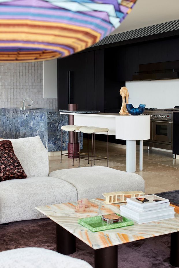 A sophisticated open-plan living and kitchen interior. The foreground features a large cream boucle sectional sofa and a low marble coffee table with dark cylindrical legs, books and decorative objects. Behind it is a contemporary kitchen with black full-height cabinetry, a blue-grey stone island and splashback, pale tiled wall, stainless-steel appliances, and a white peninsula with curved detailing. A large colourful striped sculptural ceiling light dominates the upper portion of the image.