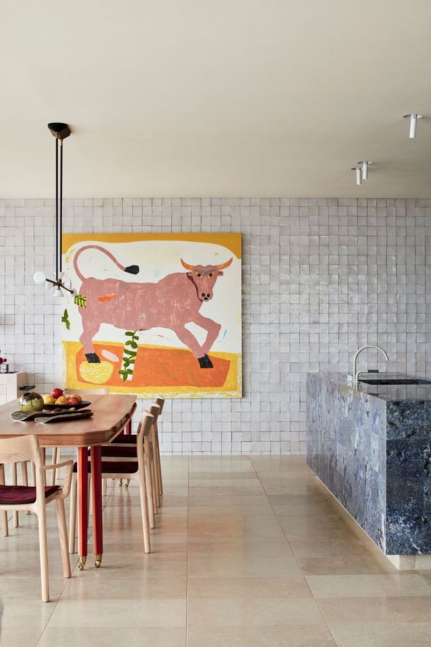 A bright contemporary kitchen and dining area with a large blue-grey stone island, pale tiled walls, and large beige floor tiles. A timber dining table with wooden chairs occupies the foreground, while a colourful bull artwork provides a bold focal point. Minimal black and white ceiling fixtures and a sculptural wall-mounted faucet complete the refined interior.