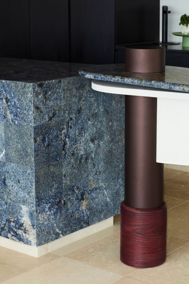 Close-up view of a highly detailed kitchen island or counter, clad in blue-grey natural stone with a richly mottled texture and rounded stone countertop edge. A white cantilevered or projecting benchtop element is supported by a sculptural cylindrical dark bronze-brown leg with a red-brown timber-like base. The background has dark matte vertical cabinetry or wall panelling, while the floor is finished in large beige tiles. The image focuses on material contrast, bespoke joinery, and a sculptural support detail.
