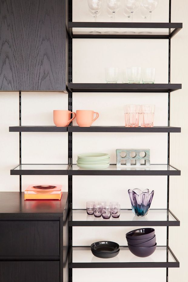 Close-up interior view of a wall-mounted open shelving system with thin black metal supports and dark shelves, displaying glassware, pastel ceramic cups and plates, decorative bowls, and a colourful glass vase. Dark built-in cabinetry is visible at the left, against a light neutral wall.
