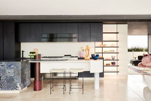 Contemporary open-plan kitchen with dark timber cabinetry, a pale stone island and integrated seating. Open shelving displays ceramics and glassware, while a bright living area is visible beyond. The space combines dark joinery with light flooring and a sculptural, design-forward fit-out.