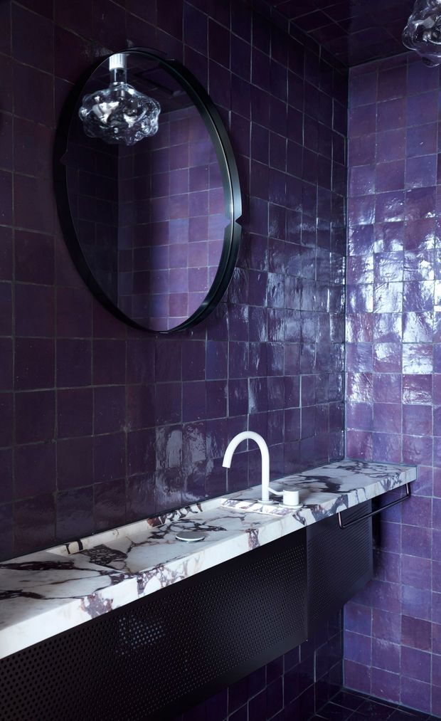 A dramatic, moody bathroom vanity clad in deep glossy purple square tiles. The wall-mounted black oval mirror reflects a sculptural pendant light, while a white-and-dark veined stone countertop supports a white curved faucet and basin fittings. The vanity base is black perforated metal, creating a luxurious, highly textured interior.