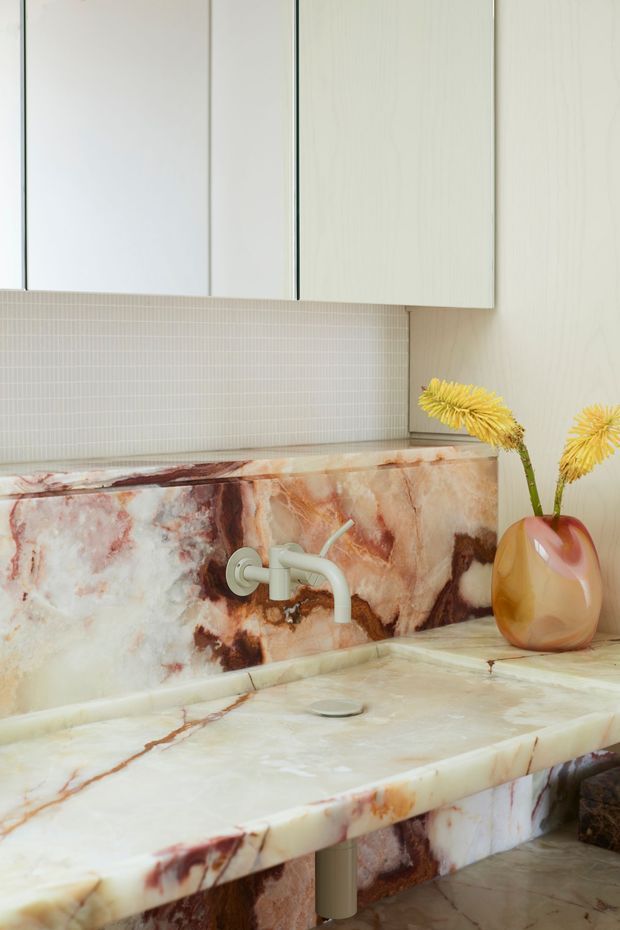 Close-up view of a refined bathroom vanity featuring dramatic veined natural stone across the benchtop, backsplash, and vanity front. A wall-mounted pale metal tap sits above the integrated basin area, with minimalist light cabinetry overhead. Small-format pale mosaic tiles line the upper backsplash, and a sculptural amber vase with dried yellow flowers adds a decorative accent. The overall design is tactile, luxurious, and softly contemporary.