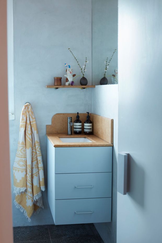 A compact, softly styled bathroom vanity nook with a pale blue wall and blue-grey floating drawer cabinet. The vanity has a warm tan textured benchtop and integrated basin, with matching tan upstands, black tapware and soap dispensers. A narrow timber shelf above holds small decorative objects and vases, while a large mirror reflects the shelf and decor. A patterned mustard and cream towel hangs beside the vanity, and dark stone-look flooring anchors the space.