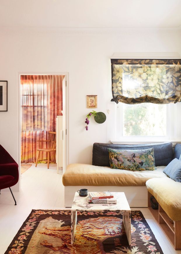 A bright, eclectic living room with a cream-painted interior, large window dressed with a dark botanical Roman blind, and a low upholstered sectional sofa in muted blue and mustard tones. A patterned vintage rug anchors a small marble-topped coffee table, while a red sculptural chair, floral cushions, framed artwork, and hanging plant add layered colour and character. An adjoining doorway reveals a smaller sitting or dining area with warm striped curtains and a timber chair.