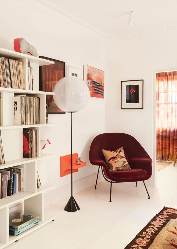 A bright, curated living room corner with white built-in shelving filled with vinyl records, a turntable, framed artwork, and a sculptural red wall shelf. A burgundy upholstered lounge chair with a patterned cushion sits beside a large spherical floor lamp. White walls and flooring create a clean backdrop, while a patterned rug and sheer rust-coloured curtains add warmth. A doorway reveals a softly lit adjoining area.