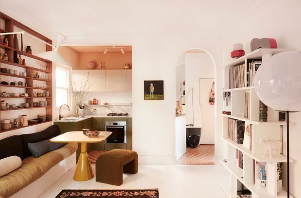 Warm, eclectic open-plan living area with a compact kitchen at the rear. The room features extensive built-in timber shelving filled with books and objects, a curved upholstered sofa, sculptural gold-toned coffee table, lounge chair, white cabinetry, arched openings, and patterned tile flooring in the adjoining passage. The palette is predominantly white and warm wood with terracotta and muted accent tones.