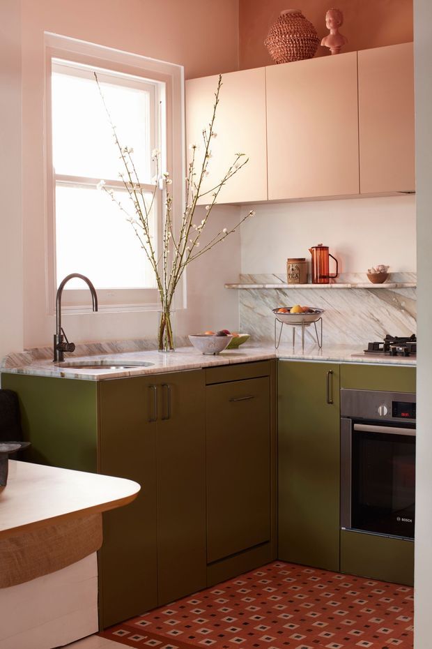A compact, warmly styled galley kitchen with olive-green lower cabinetry, pale blush-pink walls, white upper cabinets, and veined marble countertops and splashback. A stainless or dark metal gooseneck tap sits by the window, while a built-in oven and gas cooktop occupy the right side. Open shelving displays ceramics and glassware, with decorative branches and woven objects adding texture. A patterned red floor runner and a rounded dining peninsula are visible in the foreground.