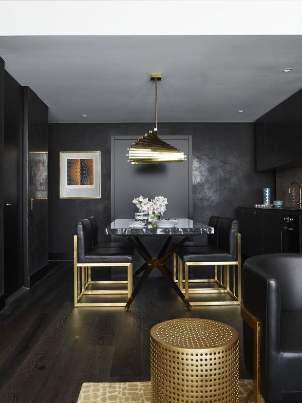 A dramatic, compact open-plan dining area with a black palette and luxurious metallic detailing. The space features a dark dining table with a marble-look top, black leather dining chairs with gold-toned metal frames, a sculptural brass pendant light, dark cabinetry and wall finishes, and dark timber flooring. A black leather sofa and perforated gold-toned side table are visible in the foreground, with framed artwork and a floral arrangement adding detail.