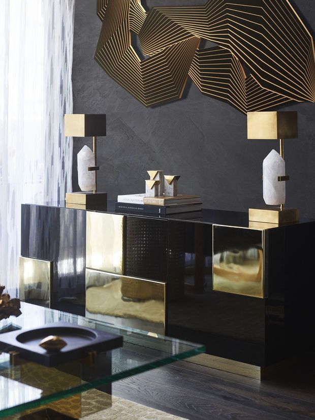 A refined, dramatic interior vignette featuring a glossy black and brass-accented sideboard against a dark textured wall. Geometric gold wall art, sculptural lamps, books, and decorative objects sit above and around the cabinetry, while a glass coffee table and dark timber flooring complete the space. Sheer curtains provide soft light from the side.
